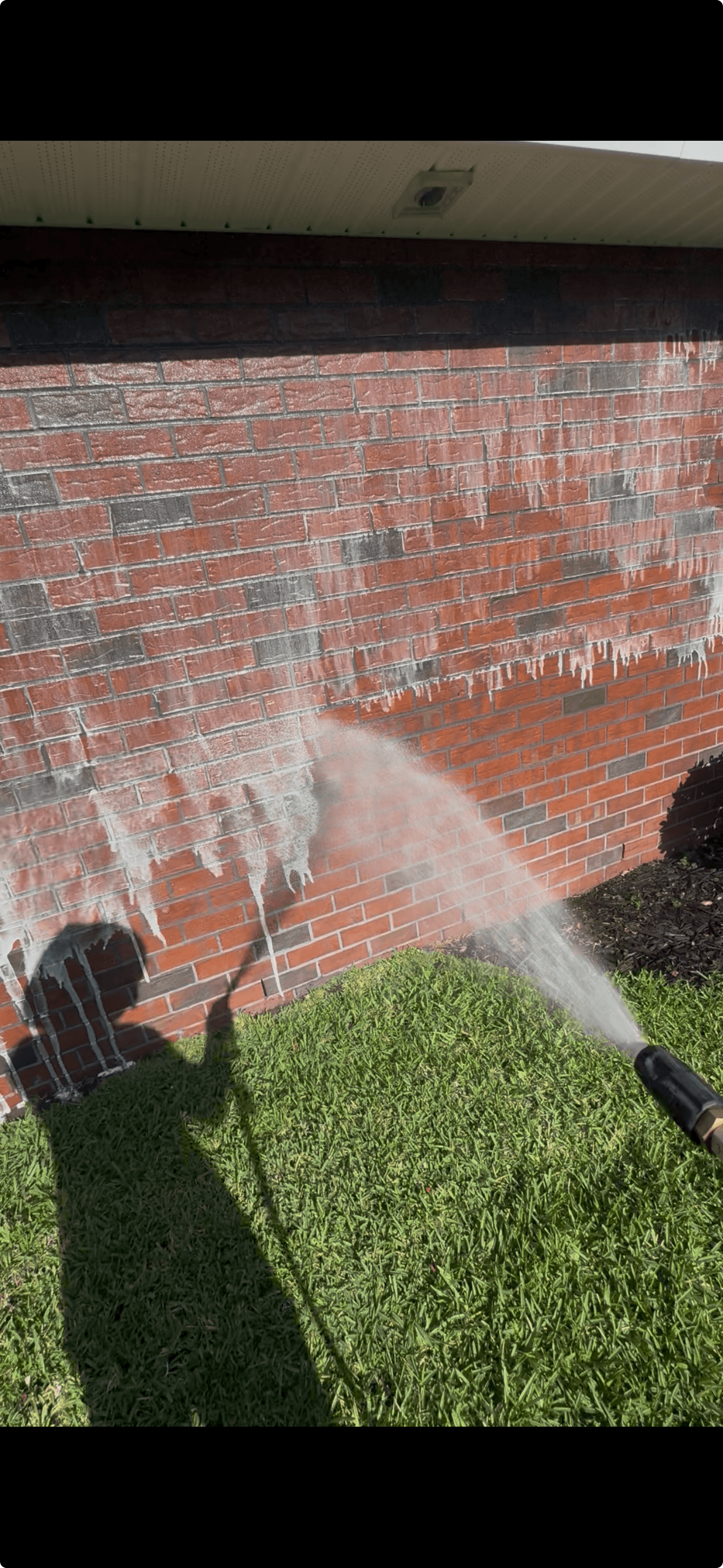 Water spray from a pressure washer cleaning white residue off a red brick wall.