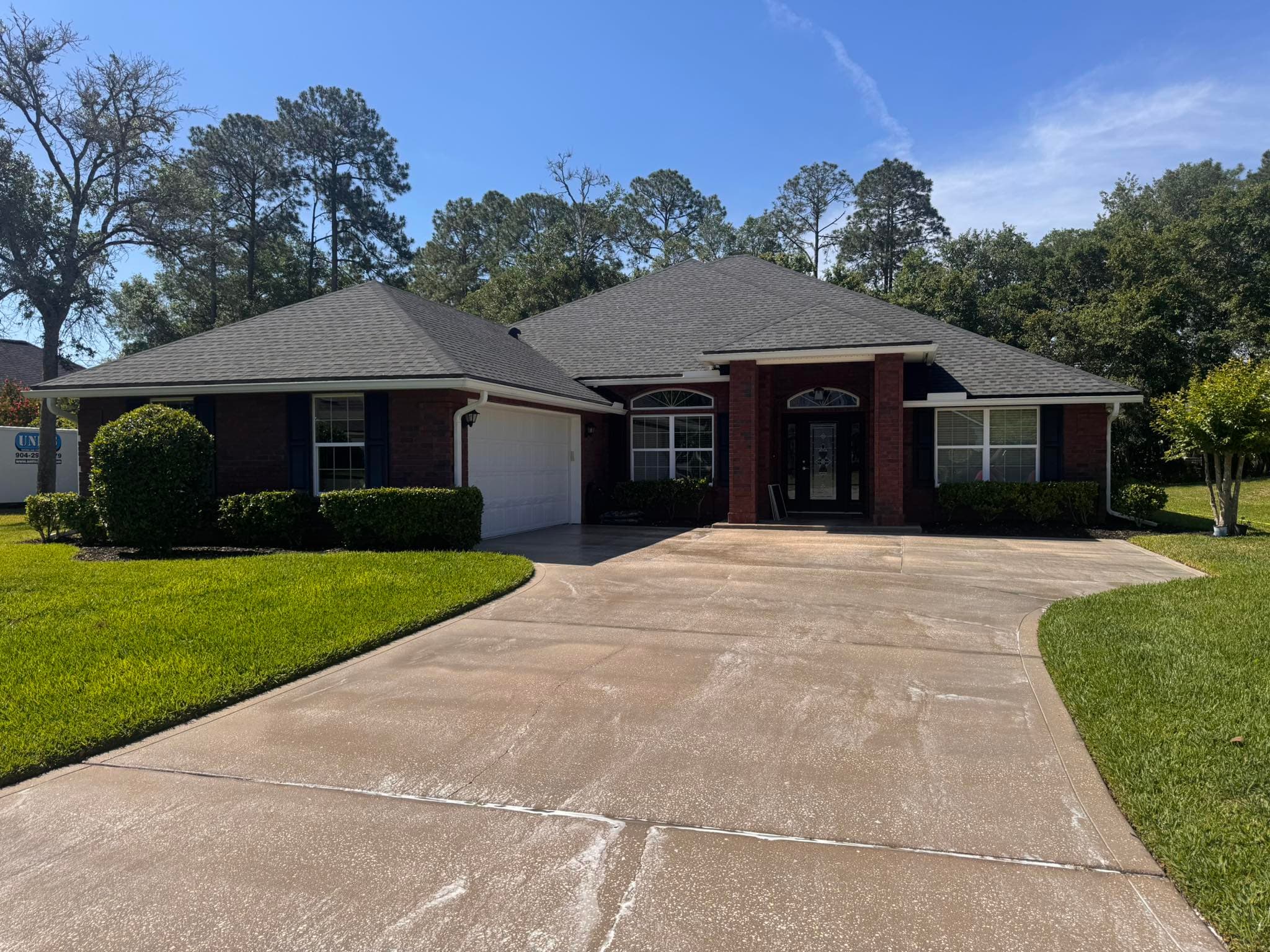 Red brick house with a wide concrete driveway, green lawn, and dark shingle roof.