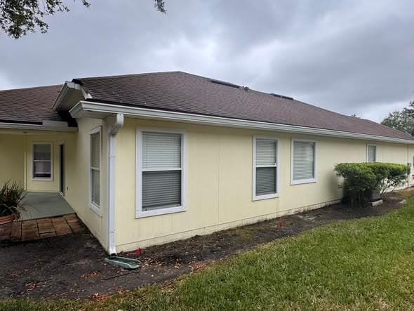 Pale yellow house with white trim, dark roof, and windows with blinds under overcast skies.