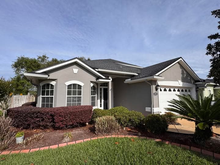 Gray single-story house with white trim, arched windows, and a garage surrounded by lush landscaping.