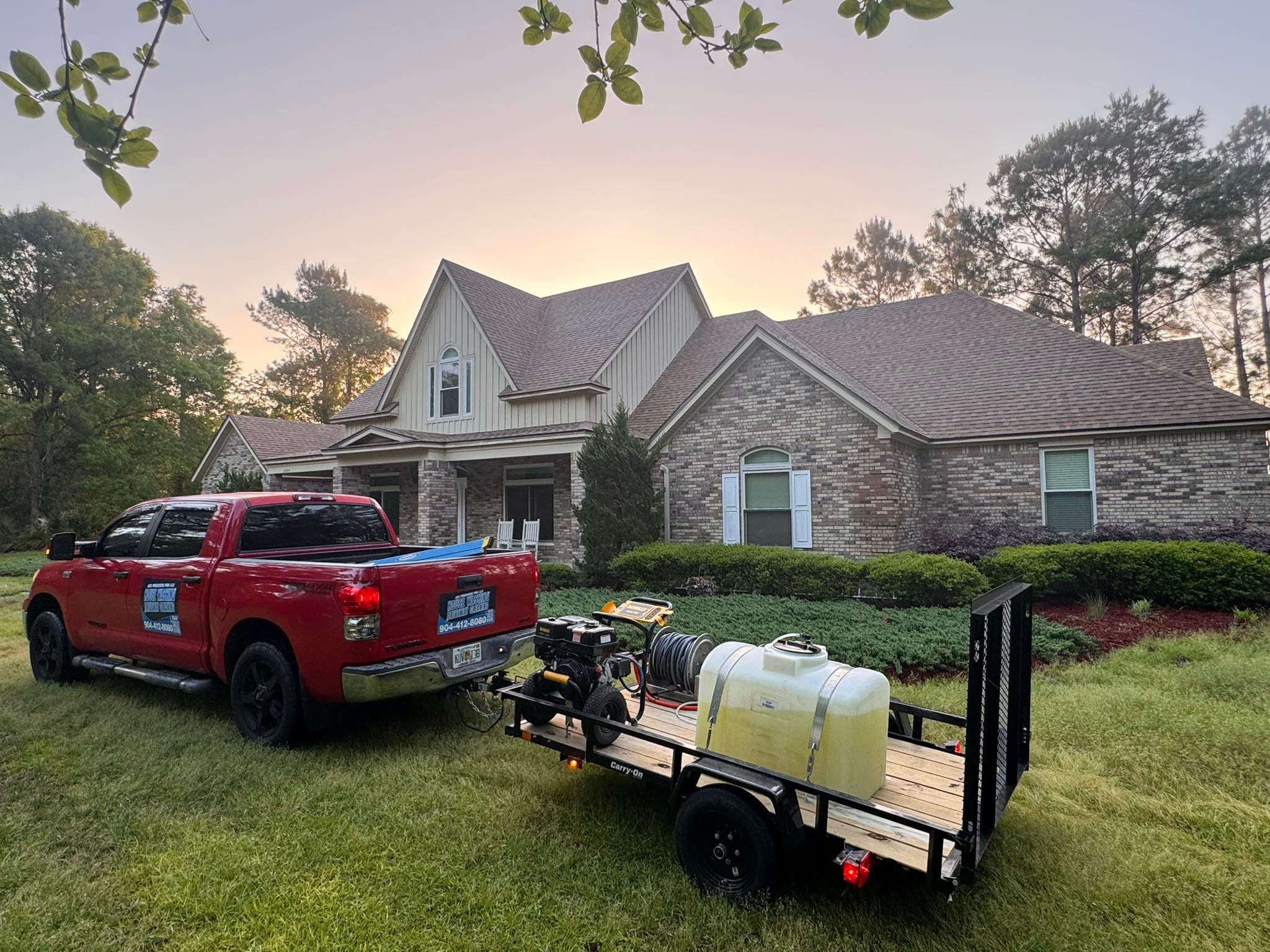 Red pickup truck towing pressure washing equipment parked in front of a large brick house at dusk.