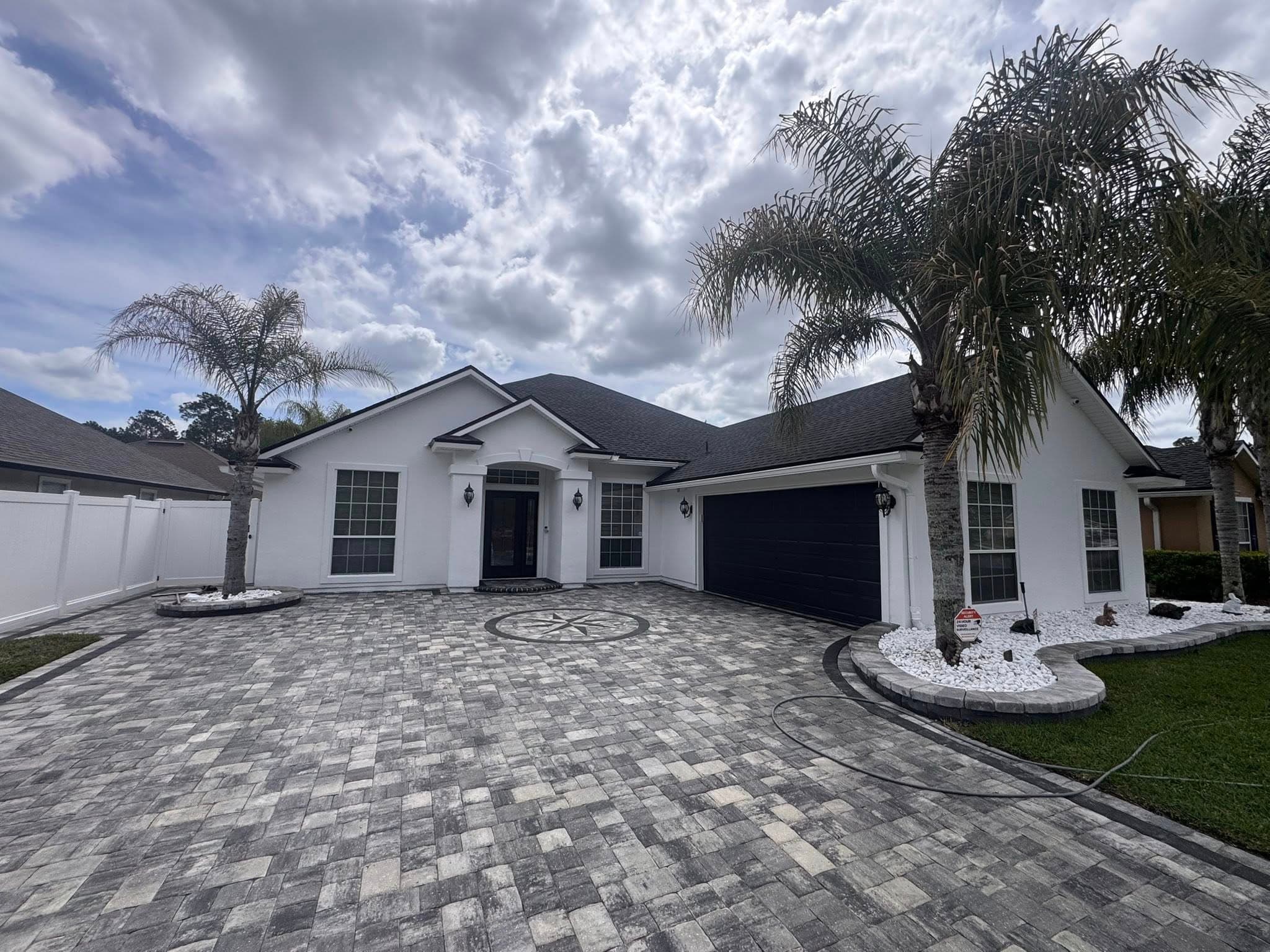 Modern white house with black accents, palm trees, and a large patterned stone driveway.